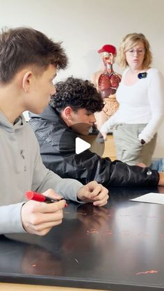 three people sitting at a table with pens and paper in front of them, one man is writing