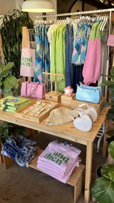 a wooden table topped with lots of clothing and bags next to potted plants in a store