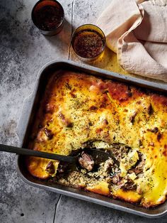 a casserole dish with meat, cheese and sauces on the side next to two glasses