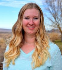 a woman with long blonde hair and blue shirt smiling at the camera while holding a cell phone in her hand