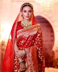 a woman in a red and gold bridal outfit with jewelry on her head, standing next to a chandelier