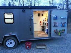 a small gray trailer with its door open and shelves on the side, sitting in gravel area next to trees