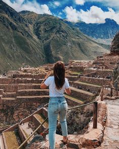 a woman standing on the edge of a cliff looking down at an ancient city with mountains in the background