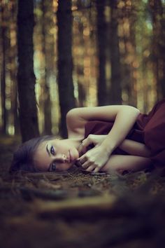 a woman laying in the woods with her hands on her chest and eyes closed, wearing a brown dress