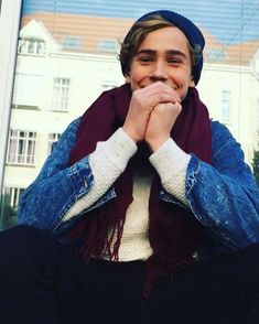 a young man sitting in front of a window with his hands on his face and wearing a scarf around his neck