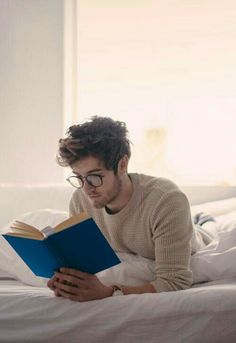 a man reading a book on top of a bed in a room with white walls