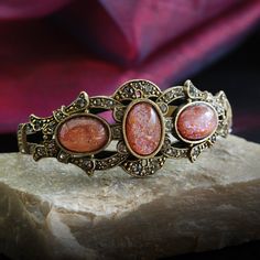 "This rich, unusually impressive bracelet is created from 90 year old stones set into our studio's original sculpture. The bracket is finely detailed and set with premium Swarovski smoke crystal. Three oval stones are vintage beauties from the 1930s which I acquired from a former exporter in Czechoslovakia. The bracket is mounted on a split shank cuff and will comfortably fit 7 to 8\" wrists. Hand burnished warm bronze finish. Entirely made in our Los Angeles studio. Choices: RUBINITE stones are Collectible Gemstone Bangle Bracelets, Elegant Bronze Bracelets For Collectors, Vintage Natural Stones Cuff Bracelet, Vintage Natural Stones Cuff Bangle, Vintage Cuff Bracelet With Natural Stones, Vintage Bangle Cuff Bracelet With Natural Stones, Vintage Cuff Bracelet With Natural Stones Gift, Vintage Bangle Bracelet With Natural Stones, Antique Gemstone Bracelet For Anniversary