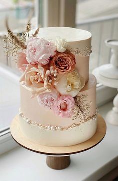 a three tiered wedding cake decorated with flowers and pearls on a wooden stand in front of a window