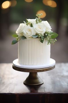 a white wedding cake with flowers on top is sitting on a wooden table in front of some lights