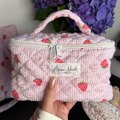 a hand holding a pink and white bag with strawberries on the front, next to purple flowers