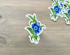 two stickers with blue flowers and green leaves on them sitting on a wooden surface