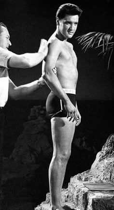 a man standing next to another man in swim trunks on top of a rocky cliff