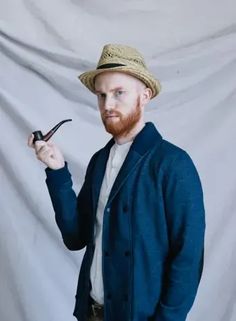 a man with a beard wearing a hat and holding a pipe in his right hand