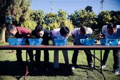 four men are standing at a long table with blue buckets on it and one man is bending over