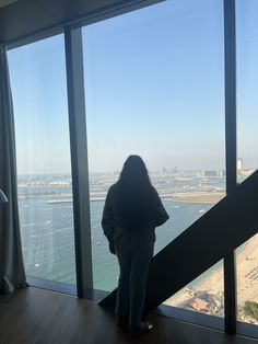 a woman standing in front of a large window looking out at the water and buildings