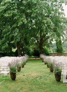 an outdoor ceremony set up with white chairs and lavender flowers on the aisle, surrounded by trees