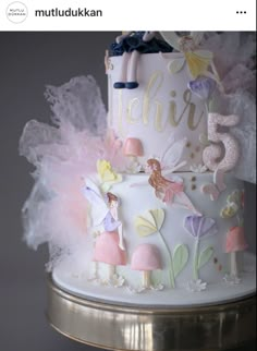 a white and pink cake with fairy figures on it's side, sitting on top of a gold stand