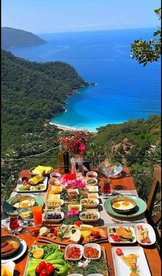 an outdoor dining table with food and drinks on it overlooking the ocean, surrounded by greenery