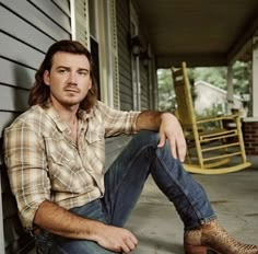 a man sitting on the side of a building next to a rocking chair and porch