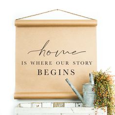 a sign that says home is where our story begins hanging on a wall next to a potted plant