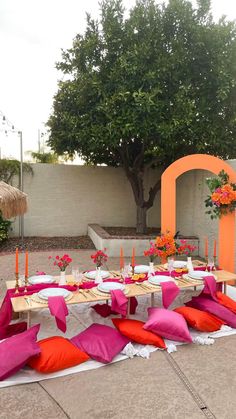 an outdoor table set up with pink and orange pillows, place settings and flowers on it