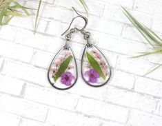 the earrings are decorated with purple flowers and green leafy leaves, on a white brick wall