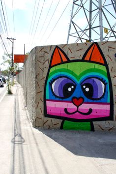 a colorful cat painted on the side of a wall next to a street with power lines