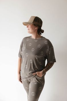 A person with a tattoo on their right forearm stands against a plain light background, wearing breathable Short Sleeve Women's Bamboo Pajamas by forever french baby in the Sage Yin Yang pattern and a tan baseball cap. They have their left hand in their pocket and are looking to the side. Maternity Pajamas, French Baby, Toddler Pajamas, Pajama Romper, Bamboo Pajamas, Bamboo Clothing, Matching Baby, Baby And Toddler, One Piece Dress