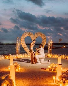 a man kneeling down next to a woman in front of a heart on the beach