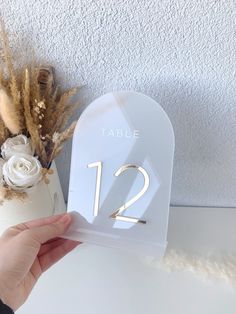 a person holding up a table number with flowers and dried grass in the background on a white surface