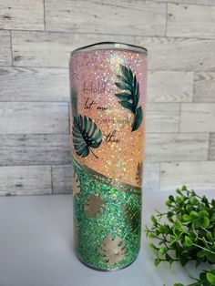 a pink and green glittered tumbler with leaves on it next to a plant