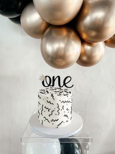 a white cake sitting on top of a table next to gold and black balloons hanging from the ceiling