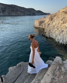 a woman in a white dress is standing on rocks near the water and looking at something