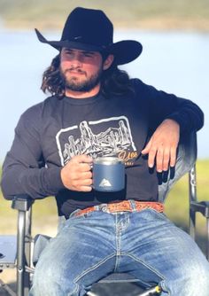 a man sitting in a chair with a cowboy hat on and holding a coffee mug