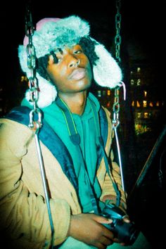 a young man wearing a hat and holding a camera in front of his face while sitting on a swing