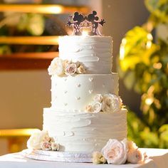 a wedding cake with white frosting and pink flowers on the top is decorated with mickey mouse figurines