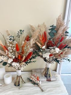 two vases filled with flowers and feathers on a white blanket next to a candle