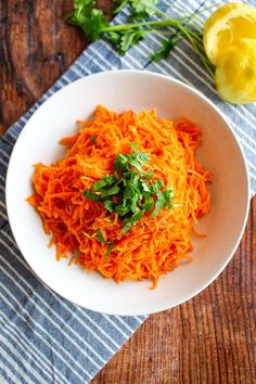 a white bowl filled with shredded carrots on top of a wooden table next to lemon wedges