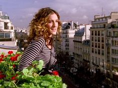 a woman standing on top of a balcony next to flowers and buildings in the background