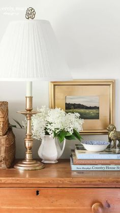 a white vase with flowers on top of a wooden dresser next to a lamp and books