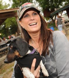 a woman holding a small dog in her arms and smiling at the camera while wearing a hat