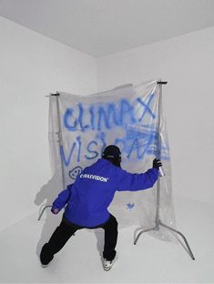 a man in blue jacket standing next to a white wall with graffiti on it and holding ski poles