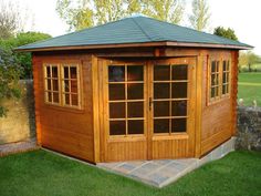 a small wooden shed with windows on the side and grass in front of it, next to a stone wall