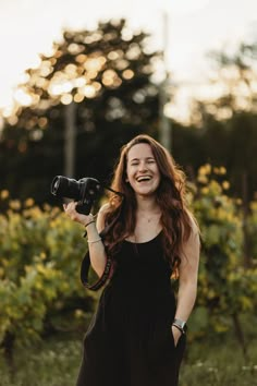 a woman holding a camera and smiling in front of some bushes with trees behind her