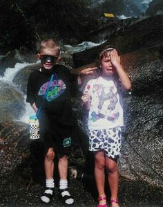 two young children standing next to each other near a river and rocks with water rushing behind them