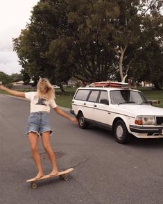 a woman riding a skateboard down a street next to a white car and trees