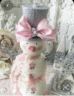 a snowman with a pink hat and bow on it's head sitting next to flowers