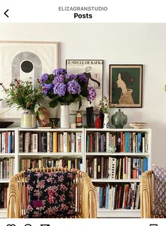 a book shelf filled with lots of books next to two chairs and a vase full of flowers