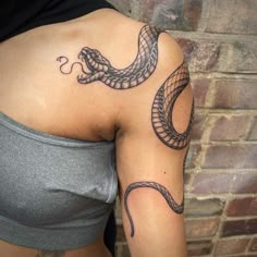 a woman with a snake tattoo on her arm and shoulder is standing in front of a brick wall