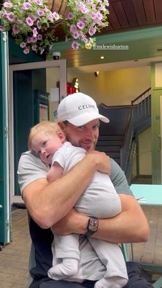 a man holding a baby in his arms while sitting on a bench with flowers hanging from the ceiling